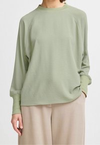 Maglione verde chiaro a maniche lunghe con scollatura rotonda, maniche raglan e tessuto testurizzato. Abbinato a pantaloni beige a gamba larga.