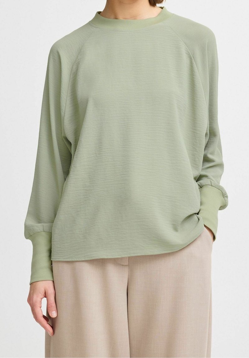 Maglione verde chiaro a maniche lunghe con scollatura rotonda, maniche raglan e tessuto testurizzato. Abbinato a pantaloni beige a gamba larga.