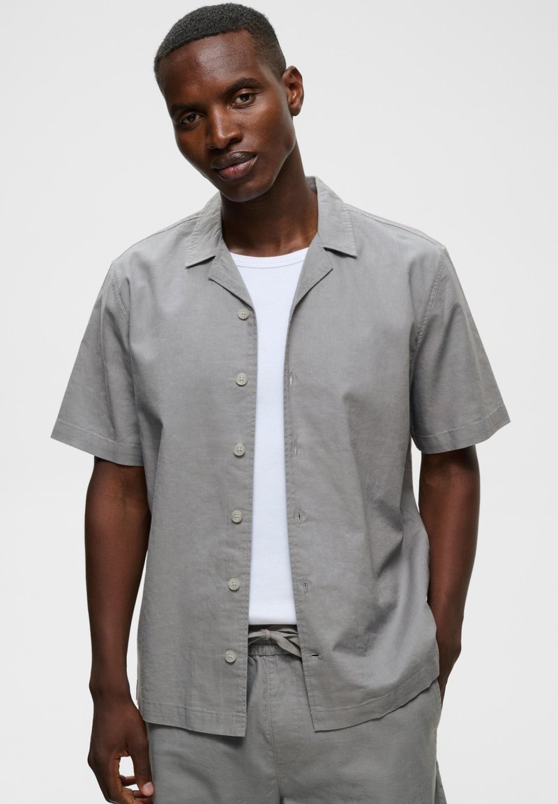 Homme aux cheveux courts portant une chemise à manches courtes gris clair boutonnée sur un t-shirt blanc et un pantalon gris assorti, debout devant un fond uni.