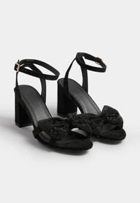 Black suede high-heel sandals with ankle straps, featuring a bow detail adorned with black beads and a square block heel.