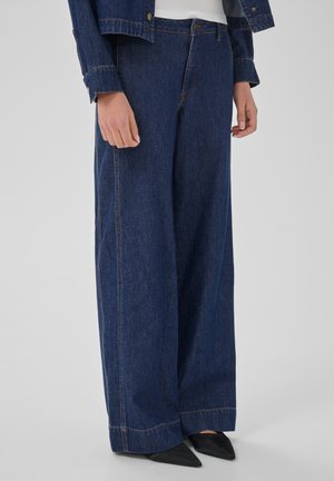 Personne portant un jean large bleu foncé taille haute et des chaussures noires pointues, vue de la taille aux pieds sur fond gris.