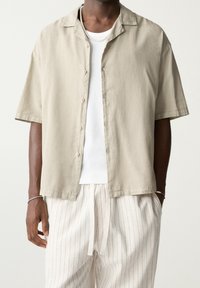 Personne à la peau foncée portant une chemise à manches courtes beige boutonnée sur un débardeur en coton blanc à côtes, un pantalon de survêtement blanc à rayures avec cordon de serrage, et des bijoux en argent.