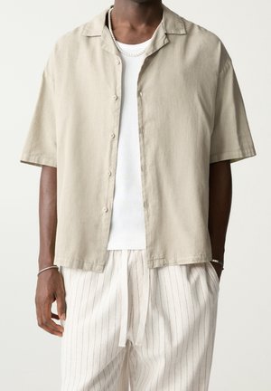 Personne à la peau foncée portant une chemise à manches courtes beige boutonnée sur un débardeur en coton blanc à côtes, un pantalon de survêtement blanc à rayures avec cordon de serrage, et des bijoux en argent.