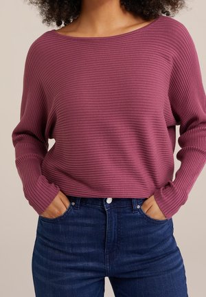 Nainen pukeutuneena pitkähihainen resorineulottu neule violettimauve värissä, leveällä pääntiellä ja tummansiniset korkeavyötäröiset farkut, kädet taskuissa.