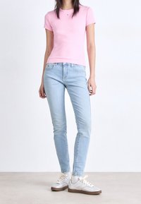 Ljust rosa, åtsittande t-shirt, blå smala jeans och silvriga sneakers med ljusa snören. Bakgrunden är enkel och neutral. Inga ytterligare detaljer.