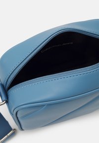 Borsa a tracolla in pelle sintetica blu, di forma rettangolare, con chiusura a zip e superficie liscia. Interno etichettato "Calvin Klein Jeans."