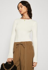 Slim fit dlouhý rukáv top v bílé tkanině, spárovaný s hnědými kalhotami na šňůrku. Modelka drží černou texturovanou tašku a předvádí vrstvené doplňky.