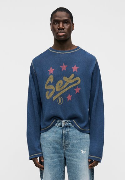 Άνδρας που φοράει μπλε πουλόβερ με τη λέξη «See» και πέντε κόκκινα αστέρια, συνδυασμένο με ανοιχτόχρωμο μπλε τζιν που έχει ένα μικρό λευκό μπαλώμα με λαγουδάκι στο αριστερό μηρό.