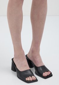 Mules en cuir noir à bout ouvert avec une forme carrée et un talon épais. Texture lisse, design minimal avec une large sangle sur le pied.