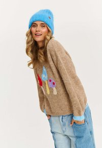 Beżowy sweter dzianinowy z kolorowym wzorem grzybów. Niebieska czapka. Miękka faktura, luźny krój. Jasnoniebieskie mankiety i swobodny styl.