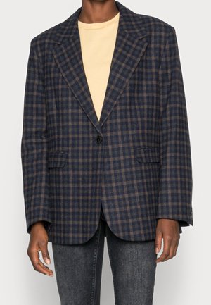 Osoba w ciemnoniebiesko-brązowej marynarce w kratę, na beżowej koszulce z okrągłym kołnierzem i ciemnoszarych dżinsach, stojąca na jednolitym tle.