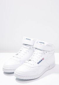 Baskets montantes en cuir blanc, dotées d'une sangle Velcro et d'une marque Reebok bleue sur le côté. Semelle texturée avec perforations.