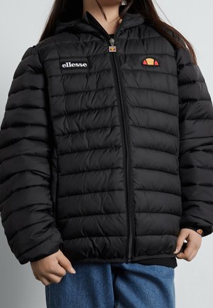 Osoba ubrana w czarną pikowaną kurtkę ellesse z logo na piersi i zamkiem błyskawicznym, w połączeniu z niebieskimi dżinsami, stojąca na jednolitym tle.
