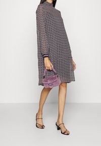 Schwarz-weiß gemustertes Kleid mit langen Ärmeln und hohem Ausschnitt, kombiniert mit einer kleinen lila Handtasche und schwarzen Riemchensandalen.