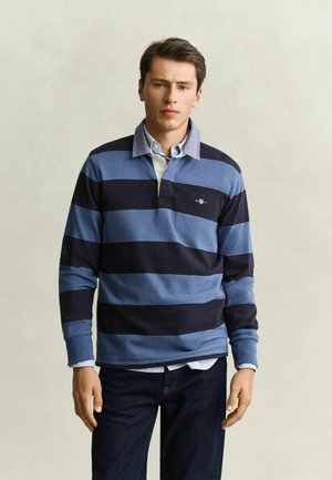 Junger Mann mit dunklen Haaren, der ein blau-schwarz gestreiftes langärmeliges Rugbyhemd und dunkle Jeans trägt und vor einem schlichten Hintergrund steht.