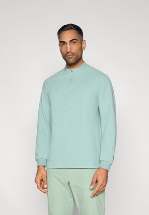 Pull-over bleu clair à col montant avec une fermeture éclair sur la moitié, fabriqué dans un tissu texturé. Manches longues et poignets côtelés, porté avec un pantalon assorti.