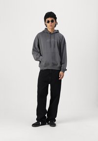 Grijze katoenen hoodie met een voorzak en trekkoorden, gecombineerd met zwarte wijdgevoerde jeans en zwarte sneakers. Accessoires zijn ronde zonnebrillen.