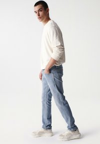 Sudadera beige chiaro, jeans azzurri e sneakers beige. I jeans hanno una vestibilità slim e dettagli di whiskering sottili.