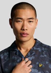 Camicia azzurra a maniche corte con un motivo testurizzato, caratterizzata da un logo giallo ricamato sul petto. Il modello mostra anelli d'argento sulle dita.