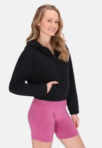 Fekete cropped kapucnis pulóver, elülső zsebből és cipzáras gallérból, rózsaszín melírozott, testhezálló rövidnadrággal párosítva, bemutatva egy laza sportos stílust.