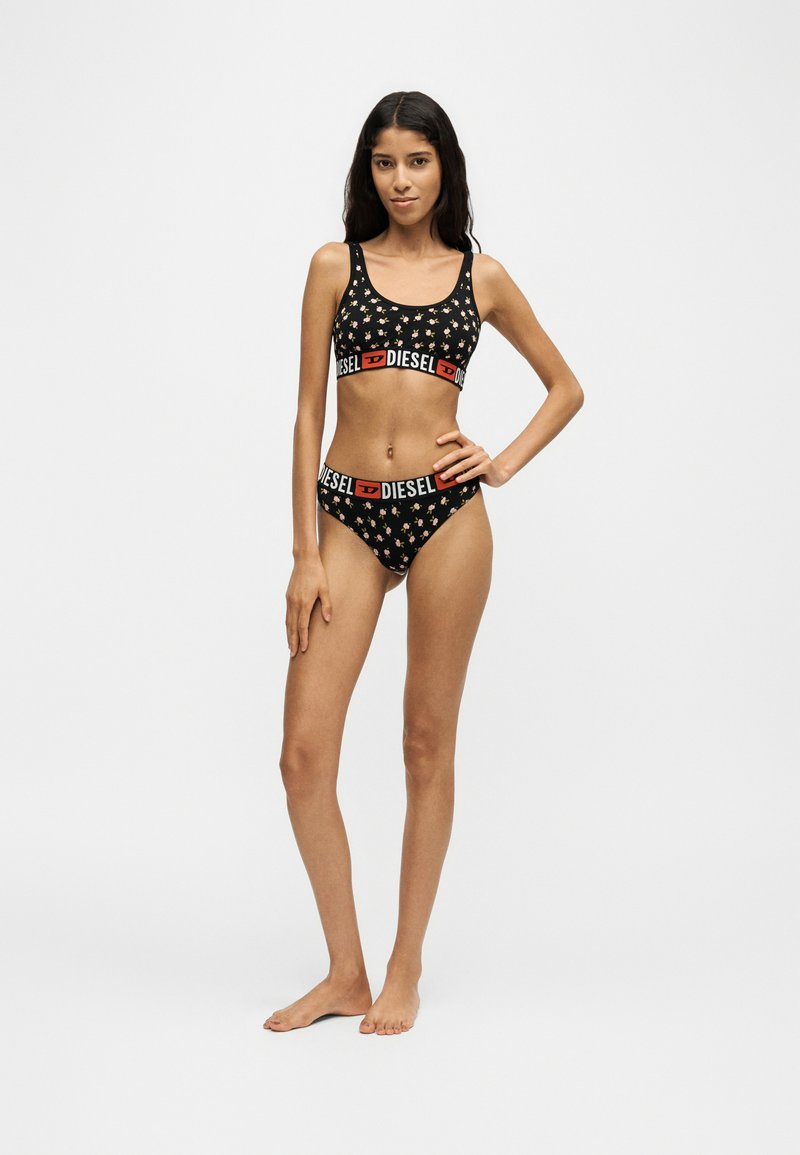 Femme debout pieds nus portant un soutien-gorge de sport noir à motifs floraux et un sous-vêtement assorti avec le logo Diesel sur les bandes élastiques.