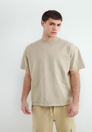 Jonge man met kort donker haar, dragend een loszittend beige T-shirt en licht beige broek, staand tegen een effen witte achtergrond.