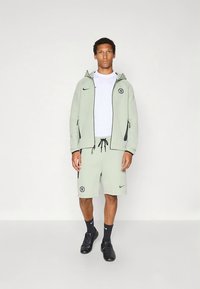 Light green hoodie with black accents, white t-shirt, matching shorts, logo on left leg, paired with black sneakers and socks.