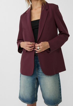 Vrouw draagt een donkerpaarse blazer over een zwart topje met lichtblauwe denim knielange shorts, met meerdere ringen aan beide handen.