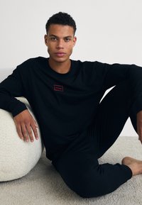Ensemble comprenant un haut à manches longues noir et un pantalon, présenté avec un petit logo rouge sur le haut. La texture semble lisse et douce sur un fond clair.