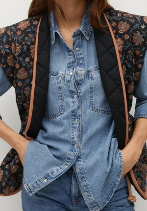 Femme portant une chemise en jean boutonnée avec des poches poitrine et un gilet matelassé à motif floral avec des bordures noires et marron.