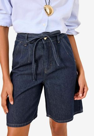 Kvinde iført mørkeblå højtaljede denimshorts med et bundet bælte, kombineret med en lyseblå opsmøgret langærmet skjorte og en guldfarvet vedhængshalskæde.