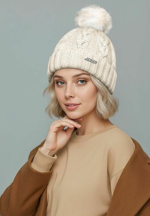 White knitted beanie with a large faux fur pom-pom, featuring a ribbed cuff and a small logo tag. Smooth texture with visible cable knit pattern.