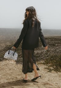 Must be mustus suur rõivaste bleiser, läbipaistev must seelik, mustad kontsad, hoides valget kotti mustade täppidega. Väliskeskkond, kus on kare maastik.