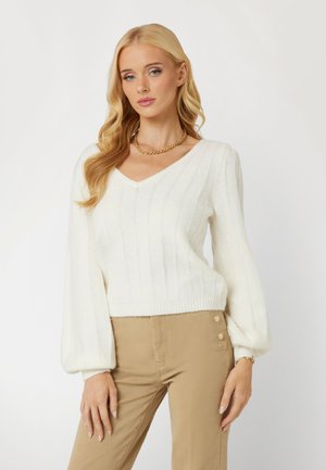 Maglione lavorato a maglia bianco con scollo a V, texture a coste e maniche a palloncino. Abbinato a pantaloni beige con dettagli di bottoni laterali.