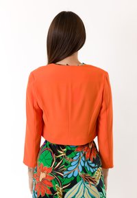 Blazer arancione accorciato con maniche lunghe, caratterizzato da una texture liscia e una cucitura posteriore. Abbinato a un vestito con motivo floreale colorato.