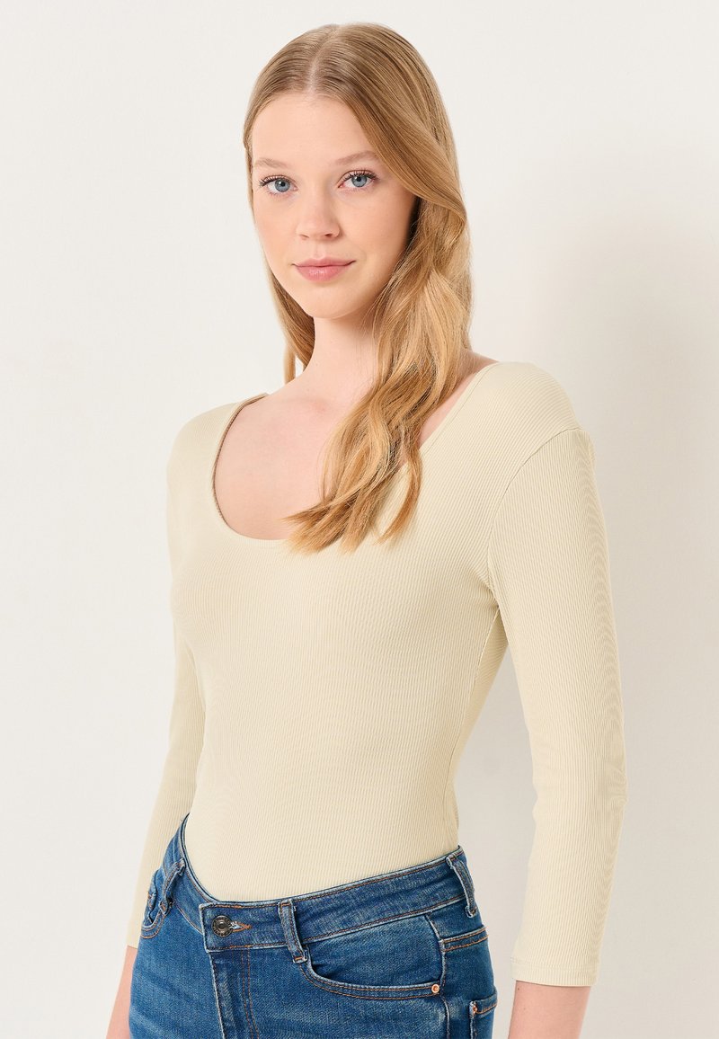 Helles beiges Rippbodsuit mit langen Ärmeln und weitem Ausschnitt, kombiniert mit enganliegenden blauen Denim-Jeans. Glatte Textur, maßgeschneiderte Passform.