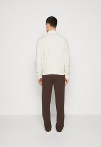 Giacca bomber beige chiaro con polsini e orlo a costine, indossata su pantaloni marroni a gamba affusolata, vista da dietro su uno sfondo bianco semplice.