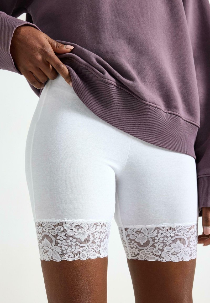 Hvide bomuldsshorts med blonder. Blonderne har blomsterprint og bølgete kanter, der fremhæver kanten. Bæres under en lilla sweatshirt.