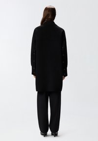 Cappotto nero oversize con colletto alto e maniche lunghe, caratterizzato da dettagli con bottoni lungo le maniche e un taglio dritto.