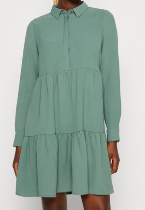 Kobieta ubrana w jasnooliwkową, długorękawową, wielowarstwową sukienkę z kołnierzykiem i zapinanym na guziki karczkiem, stojąca na jednolitym białym tle.