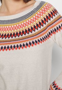 Femme portant un pull en maille beige avec des motifs géométriques colorés en orange, marine et jaune autour de l'encolure et des poignets.
