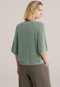 Pull en maille vert clair avec manches raglan, tissu texturé et coupe décontractée. Présenté avec un pantalon ample de couleur neutre.