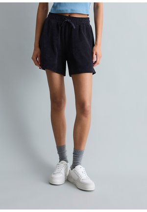 Person trägt schwarze Shorts mit Kordelzug, graue Sportsocken, weiße Turnschuhe und ein kurzes blau geripptes Oberteil vor einem einfarbigen grauen Hintergrund.