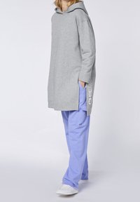 Felpa grigia con cappuccio e fessure laterali, con testo bianco sulla manica. Abbinata a pantaloni azzurri chiari e sneakers bianche.