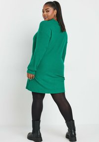 Vestido de punto verde con mangas largas, que llega a mitad del muslo, combinado con botas negras hasta el tobillo y medias negras opacas. Textura suave.