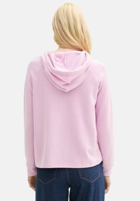 Svetloružová mikina s veľkou, mäkkou kapucňou. Má rebrované manžety a lem, s voľným strihom. Pohľad zozadu, kombinovaná s modrými džínami.