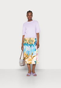 Sweater cropped em malha lilás, saia floral em camadas nas cores azul, amarelo e rosa, sandálias deslizantes em lavanda e uma mala cruzada cinza.