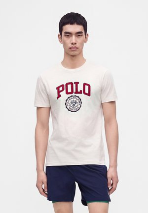 Νέος άνδρας που φοράει λευκό t-shirt Polo Ralph Lauren με κόκκινο κείμενο και σκούρα μπλε σορτς, στέκεται σε ένα απλό φωτεινό φόντο.