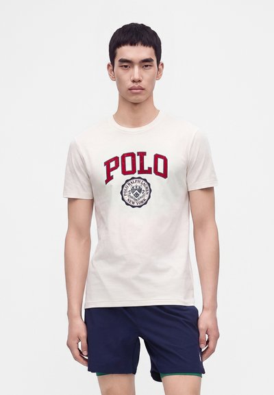 Νέος άνδρας που φοράει λευκό t-shirt Polo Ralph Lauren με κόκκινο κείμενο και σκούρα μπλε σορτς, στέκεται σε ένα απλό φωτεινό φόντο.