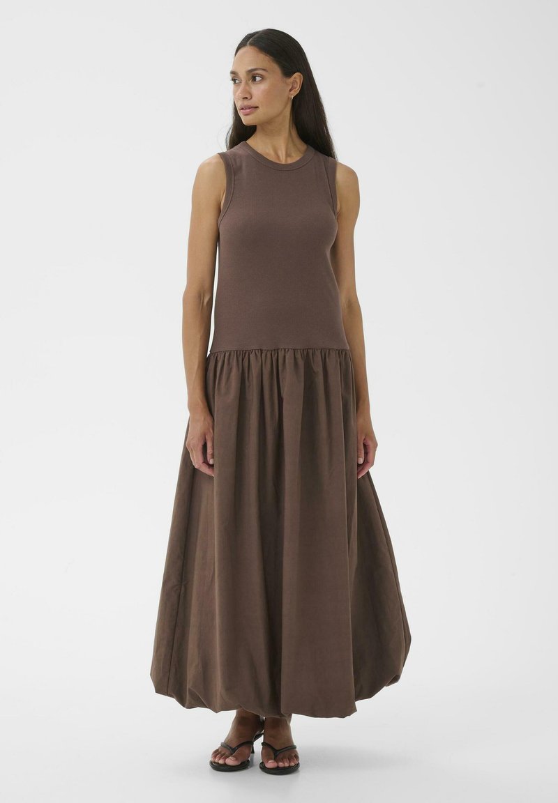 Femme aux longs cheveux foncés portant une robe maxi marron sans manches avec jupe froncée et sandales noires à brides, debout sur fond blanc.